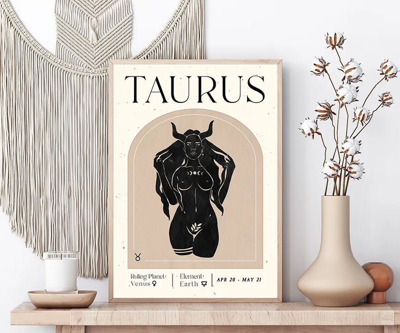 A framed Taurus astrology art print displayed on a wooden shelf, styled with a macrame wall hanging, a candle, and a vase with dried flowers.