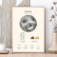 A framed astrology poster for Gemini displayed on a wooden surface next to a beige vase with dried flowers and a cream macrame wall hanging. The poster features a watercolor moon graphic, the Gemini zodiac symbol, birthstone suggestions, and planetary symbols.