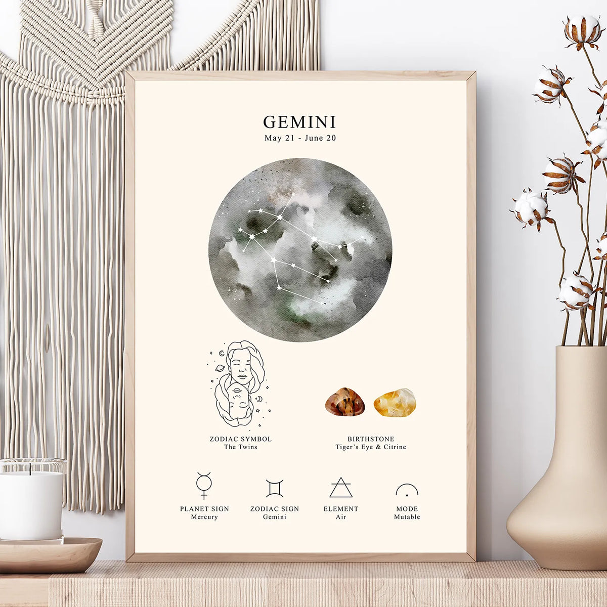 A framed astrology poster for Gemini displayed on a wooden surface next to a beige vase with dried flowers and a cream macrame wall hanging. The poster features a watercolor moon graphic, the Gemini zodiac symbol, birthstone suggestions, and planetary symbols.