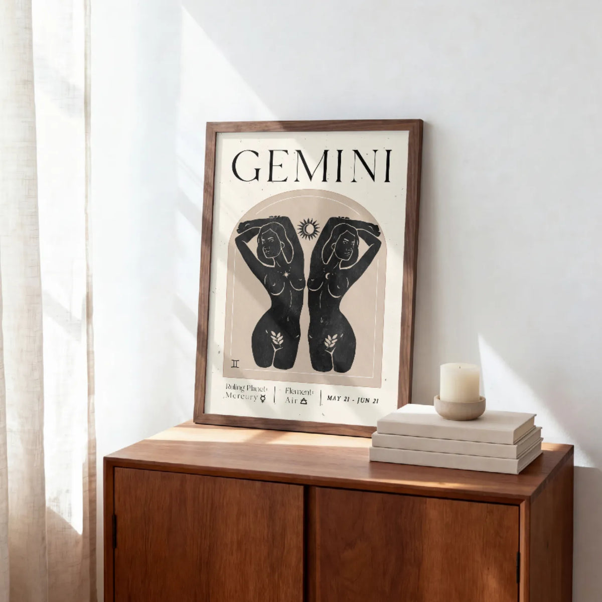 A framed art print featuring a black and beige Gemini zodiac sign illustration, styled on a wooden sideboard next to a stack of books and a candle in a bright room.