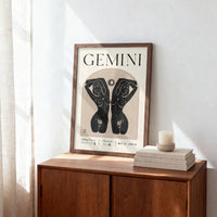 A framed art print featuring a black and beige Gemini zodiac sign illustration, styled on a wooden sideboard next to a stack of books and a candle in a bright room.