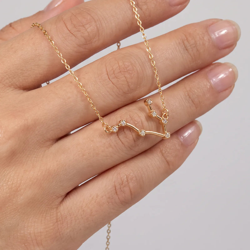 A gold necklace with a Gemini constellation crystal pendant, shown resting on a person's hand against a light gray background. 