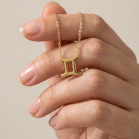 A gold necklace with a Gemini zodiac symbol pendant, held on a person's fingers against a soft beige background.