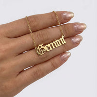 A gold nameplate necklace spelling 'Gemini' in a gothic font, shown resting on a person's hand against a light gray background.