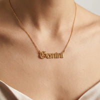 A gold nameplate necklace spelling gothic 'Gemini' worn on a model's neck, shown in a close-up shot against a neutral light gray background.