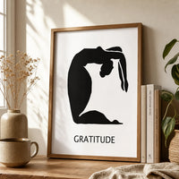 A minimalist black graphic print featuring a silhouette of a person in a graceful arching pose, with the word 'GRATITUDE' and a brown frame
