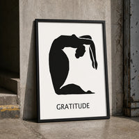 A minimalist black graphic print featuring a silhouette of a person in a graceful arching pose, with the word 'GRATITUDE' in a black frame