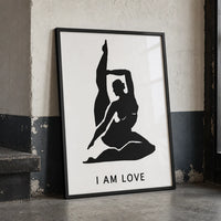 A minimalist black graphic print featuring a silhouette of a woman in a yoga pose with her leg raised, above the text 'I AM LOVE'  in a lack frame