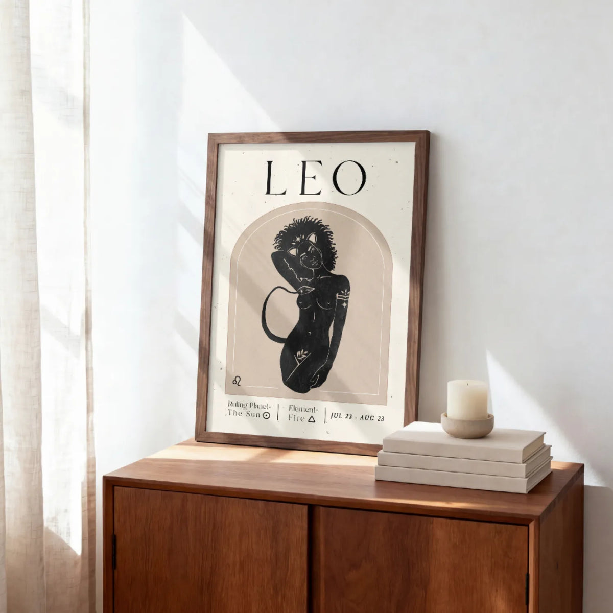 A framed art print featuring a black and beige astrological illustration of Leo with the word 'LEO' at the top, styled on a wooden cabinet next to a candle and books in a bright room.