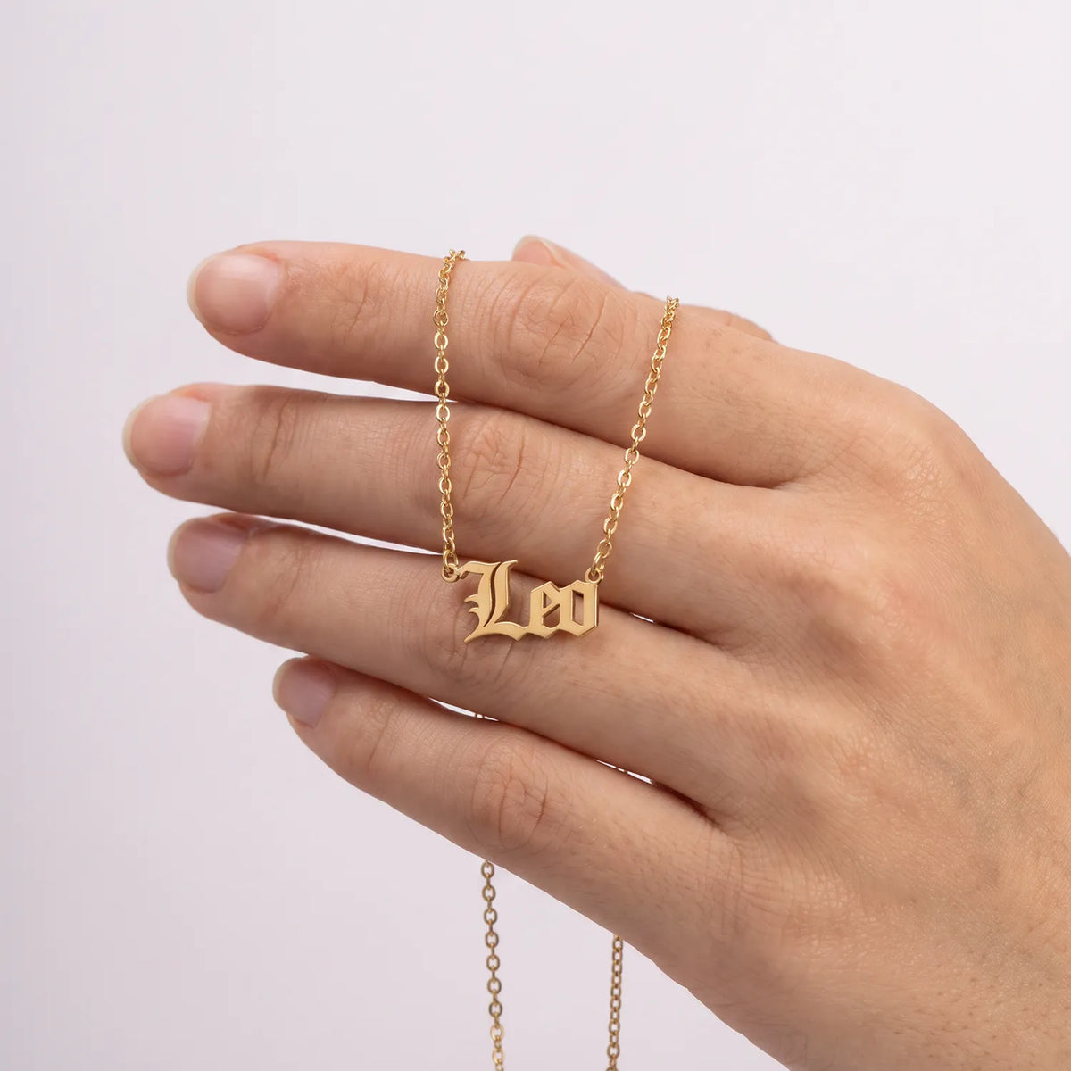 A gold chain necklace with a gothic-style 'Leo' initial pendant is shown worn on a person's finger against a plain light background.