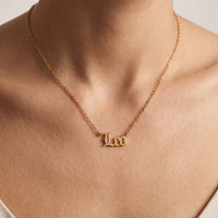 A close-up of a person's neck and chest wearing a delicate gold chain necklace with a small gothic-style 'Leo' initial pendant.