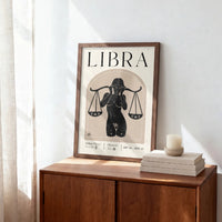 A framed Libra poster leaning on a wooden cabinet in a sunlit room, with a stack of books and a candle on top.