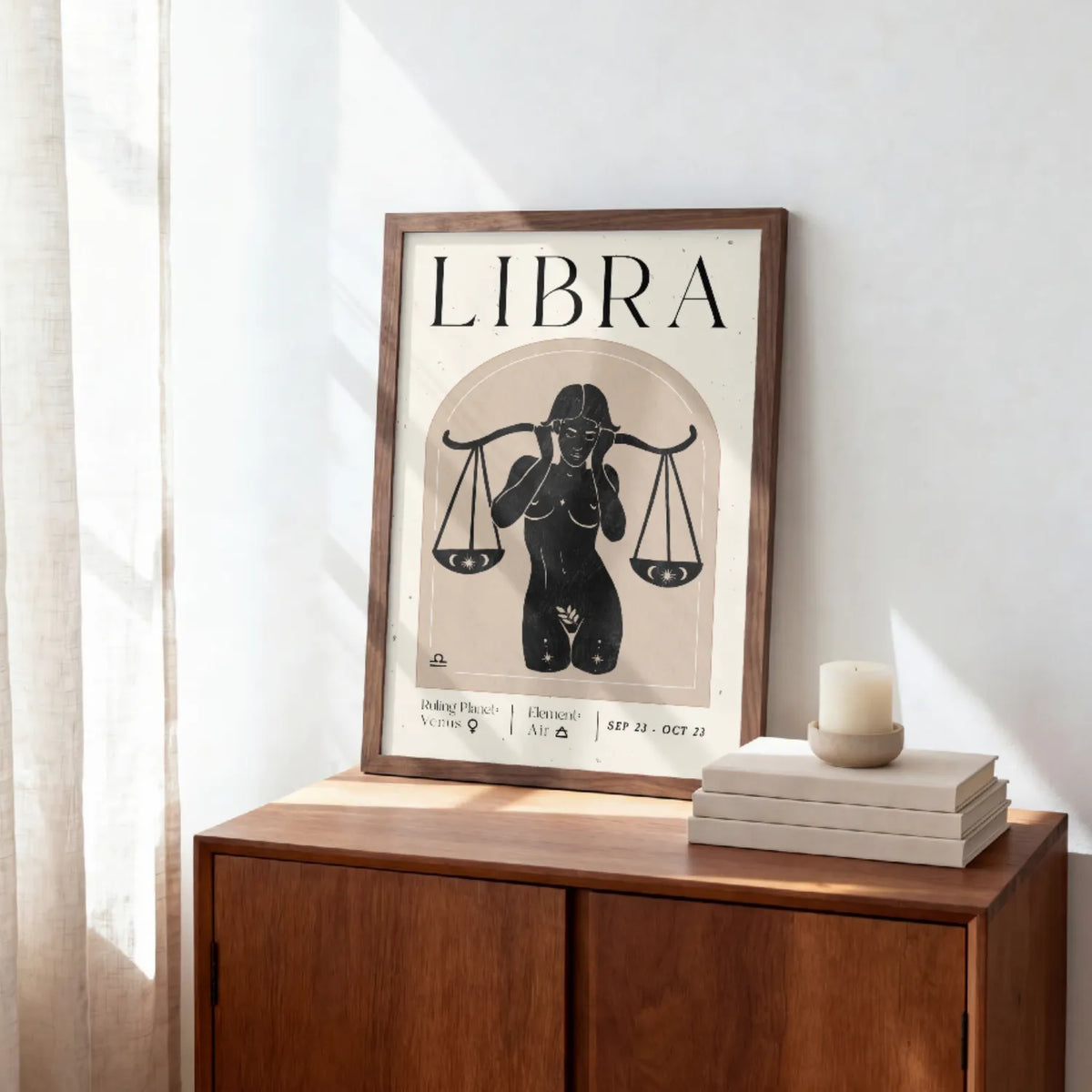 A framed Libra poster leaning on a wooden cabinet in a sunlit room, with a stack of books and a candle on top.
