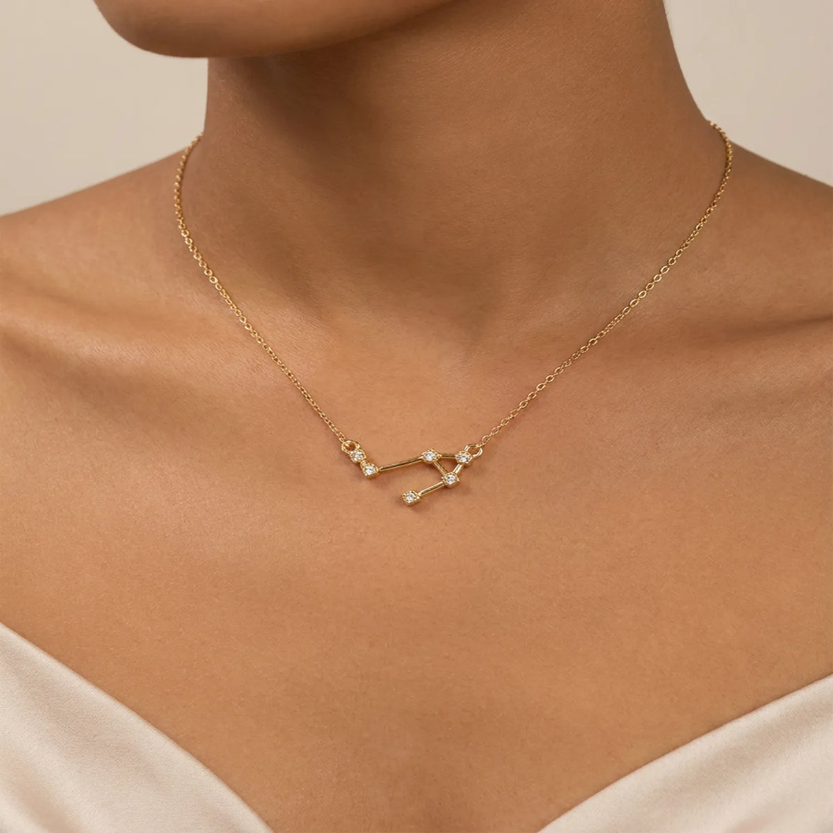 A delicate gold necklace with a libra constellation pendant, worn on the neck of a model wearing a cream-colored top.
