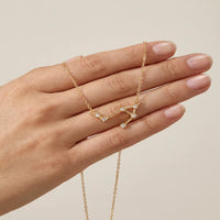 A gold necklace with a libra constellation set with small clear stones, shown resting on a person's hand against a soft beige background.