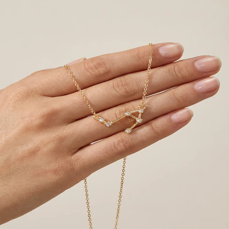 A gold necklace with a libra constellation set with small clear stones, shown resting on a person's hand against a soft beige background.