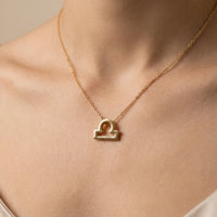 A gold necklace with a Libra zodiac sign pendant, worn on a model's neck against a soft beige background.