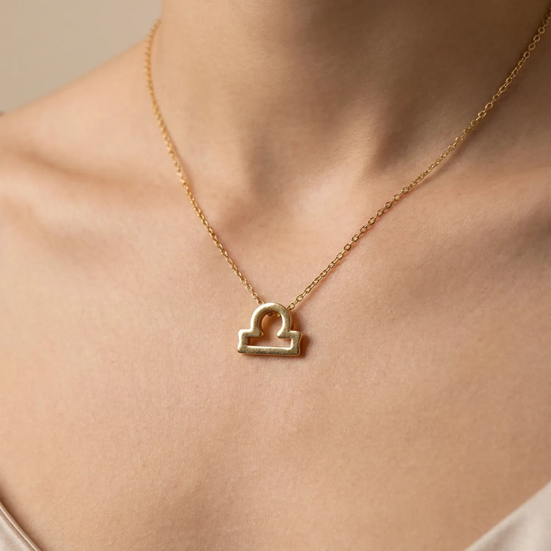 A gold necklace with a Libra zodiac sign pendant, worn on a model's neck against a soft beige background.