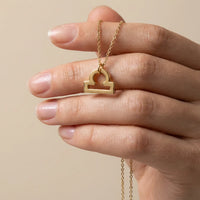 A gold necklace with a Libra zodiac sign pendant is held by a hand against a warm beige background.