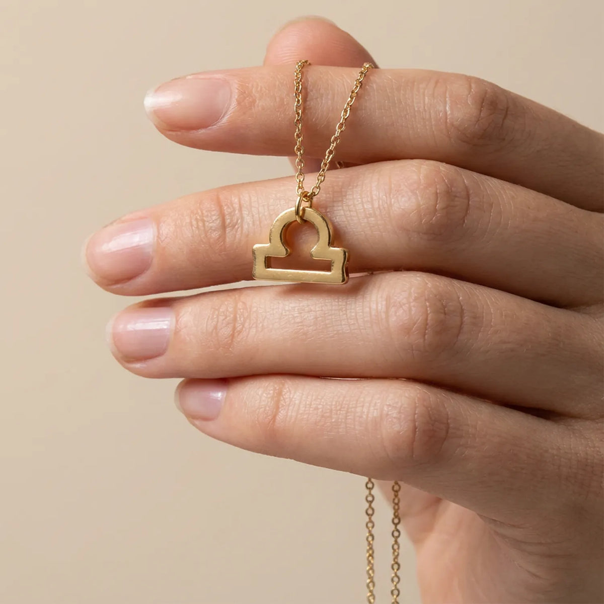 A gold necklace with a Libra zodiac sign pendant is held by a hand against a warm beige background.