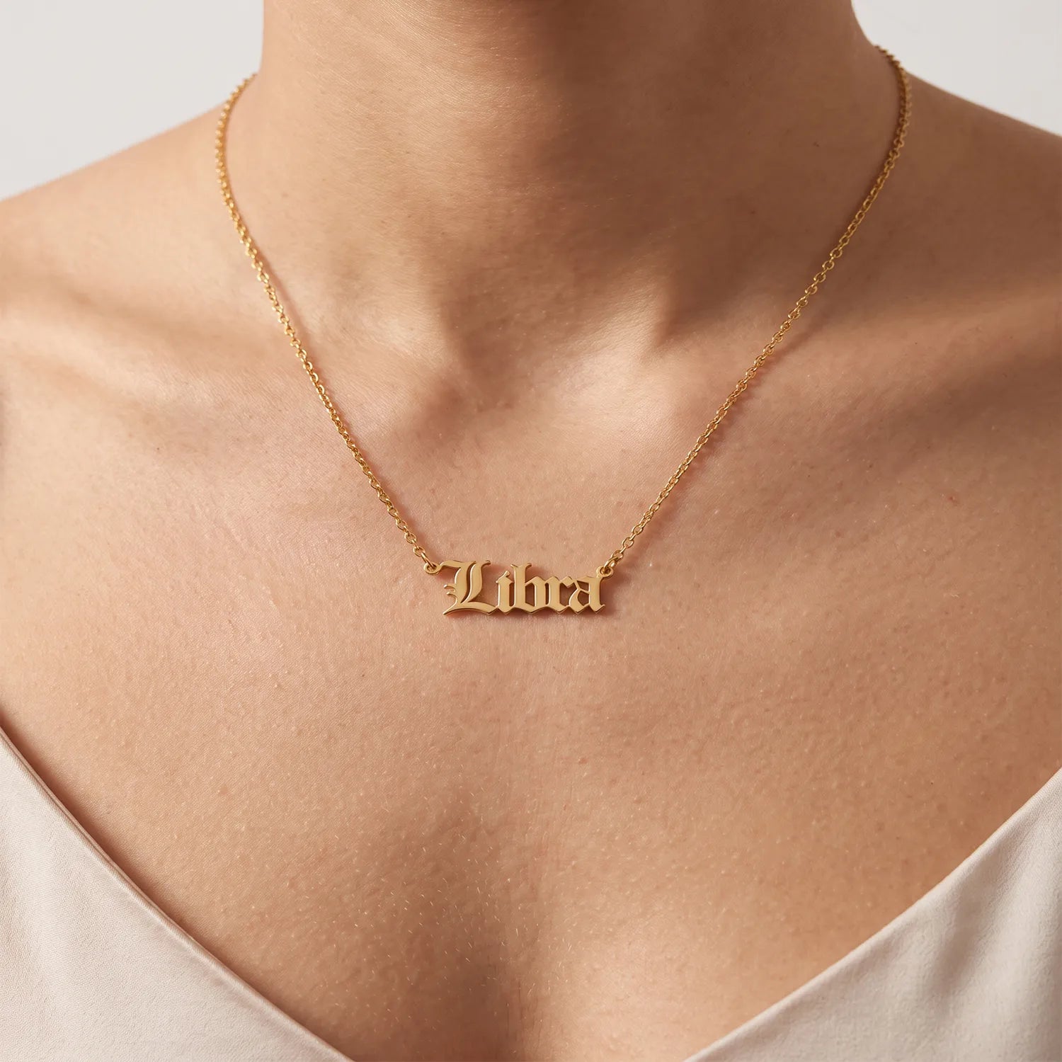 A gold nameplate necklace spelling Libra in a gothic font worn on a model's neck, shown in a close-up shot with a neutral background.