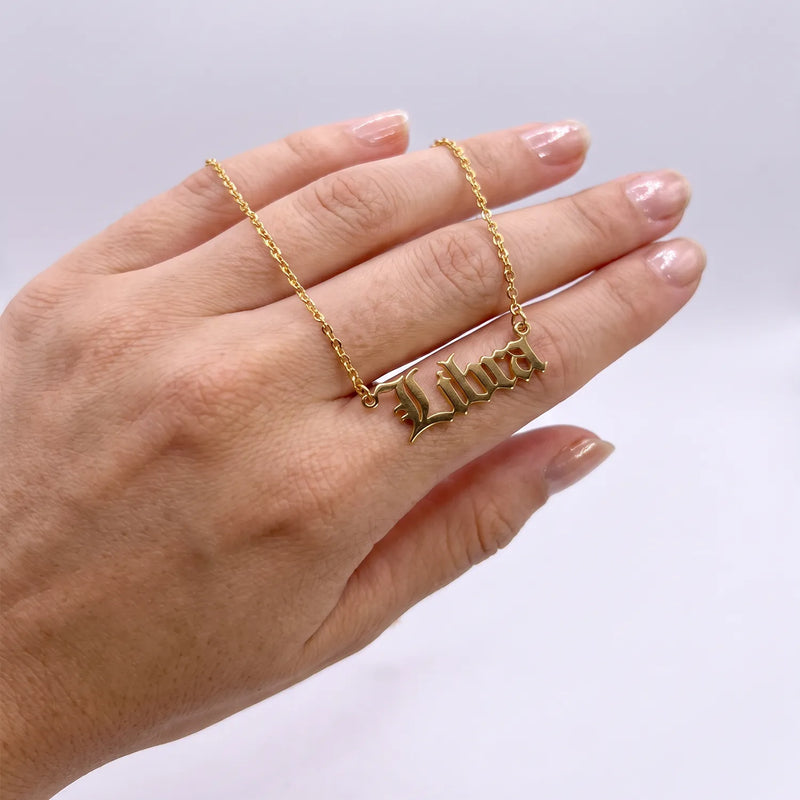 A gold nameplate necklace spelling Libra in a gothic font, worn on a person's ring finger against a light gray background.