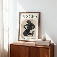 A framed Pisces zodiac art print with a black illustration of a woman holding a pitcher, styled on a wooden sideboard next to a candle and books.
