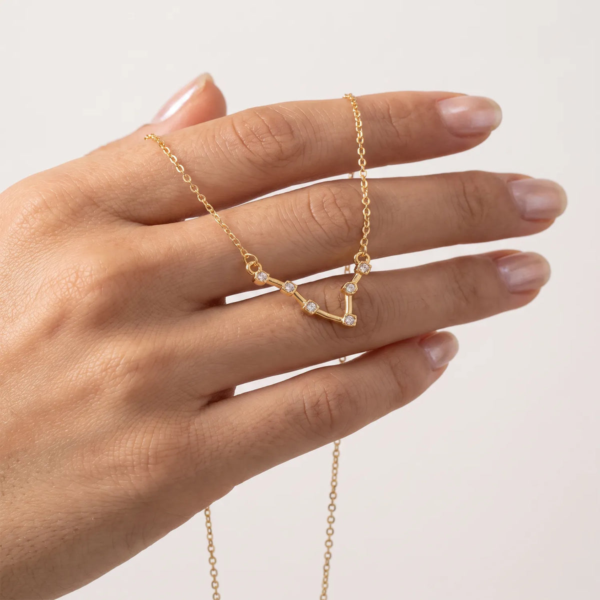 A delicate gold necklace with a small pisces constellation pendant is draped across a person's hand against a clean, light off-white background.
