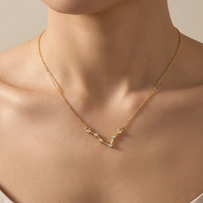 A delicate gold necklace with a small pisces constellation pendant featuring tiny clear stones, shown worn on a model's neck against a soft beige background.