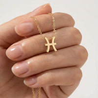 A gold chain necklace with a Pisces Zodiac symbol pendant held by a hand with neatly manicured nails against a clean white background.