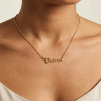 A close-up of a model's neck and chest wearing a delicate gold chain necklace with a small gothic-style script pendant spelling Pisces