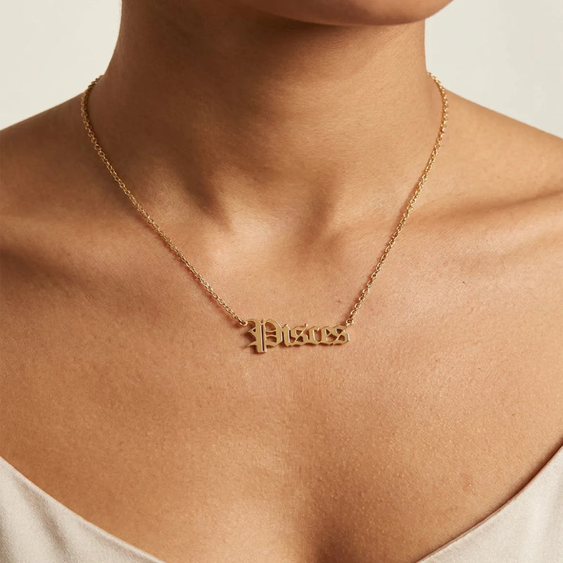 A close-up of a model's neck and chest wearing a delicate gold chain necklace with a small gothic-style script pendant spelling Pisces