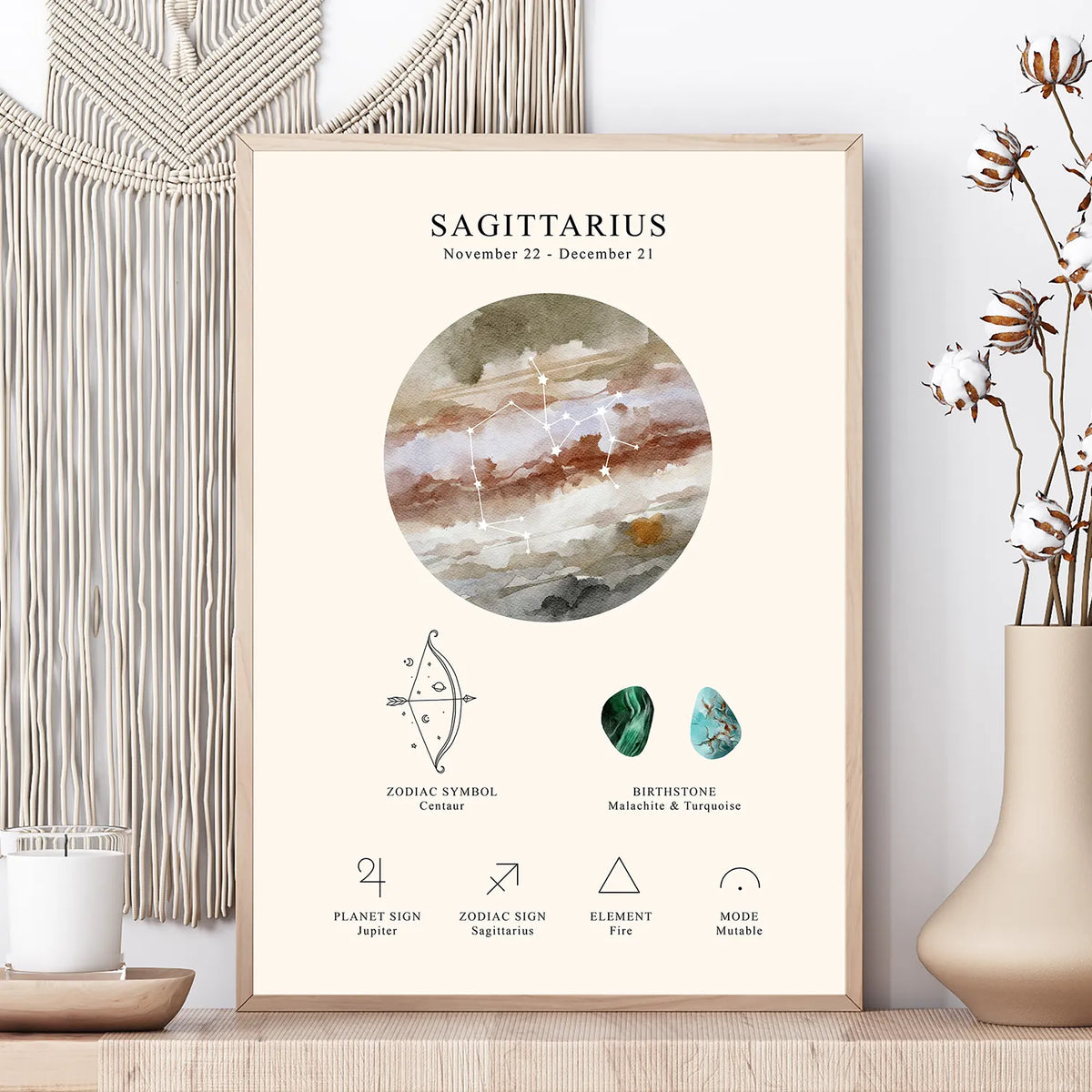 A framed Sagittarius zodiac art print styled on a wooden surface next to a macrame wall hanging and a vase with dried flowers. The print features a watercolor illustration of a brown and beige agate stone in a circular frame, with astrological details including the zodiac symbol, birthstones, and elemental associations.