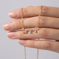 A delicate gold necklace with a sagittarius constellation in gothic script pendant featuring clear gemstones, held on a person's fingers against a soft gray background.
