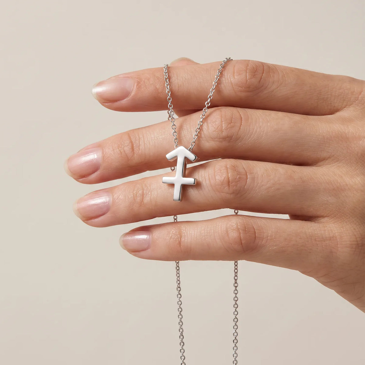 A golden necklace with a Sagittarius zodiac symbol pendant held by a hand against a solid beige background.