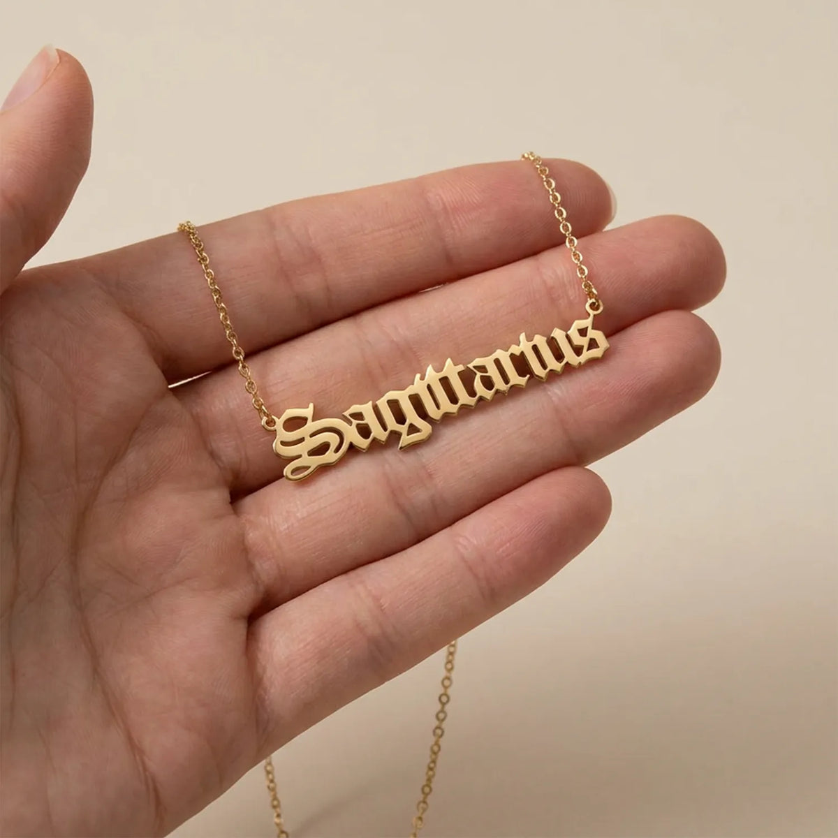 A gold nameplate necklace spelling 'Sagittarius in a gothic script held in a person's hand against a plain beige background.