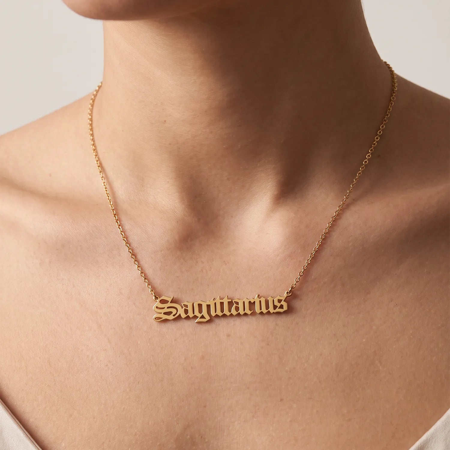 A close-up of a person's neck and upper chest wearing a delicate gold chain necklace with a nameplate pendant spelling 'Sagittarius' in a cursive, gothic-style font.