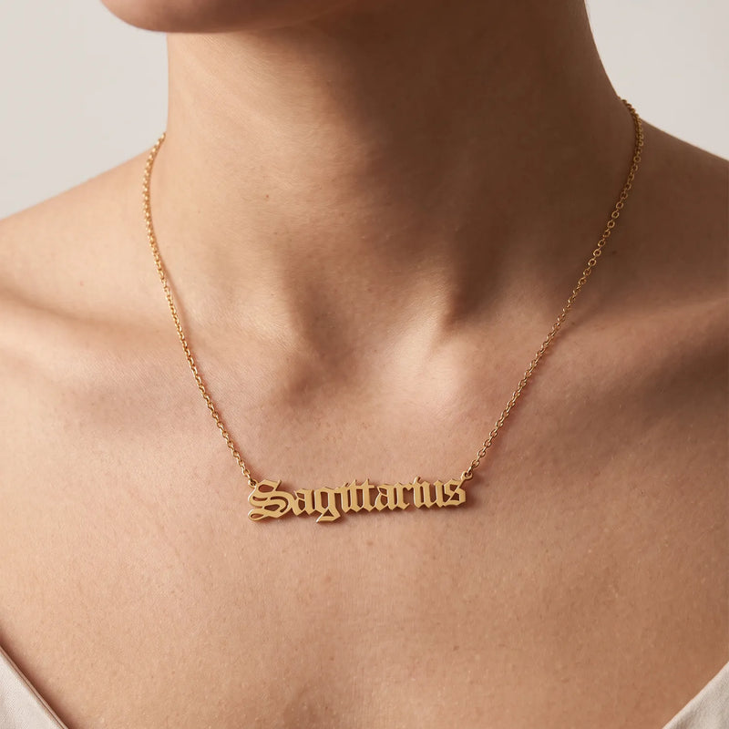 A close-up of a person's neck and upper chest wearing a delicate gold chain necklace with a nameplate pendant spelling 'Sagittarius' in a cursive, gothic-style font.