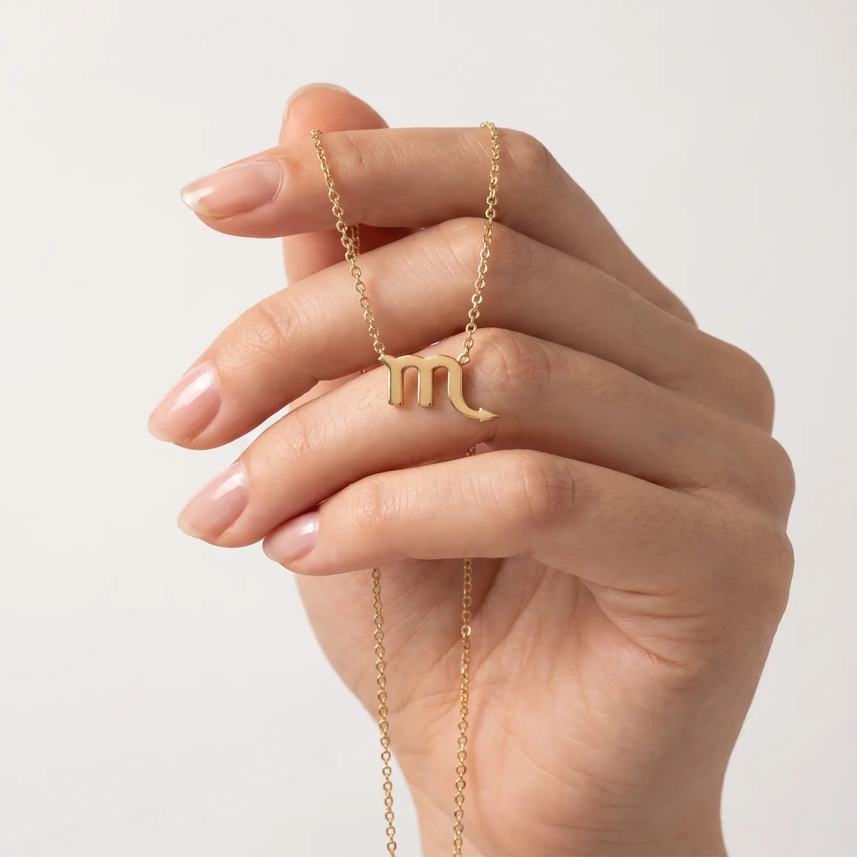 A close-up of a hand holding a delicate gold chain necklace with a small zodiac scorpio sign pendant, set against a clean, light off-white background.