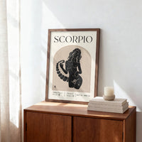 A framed art print featuring a black silhouette of a Scorpio zodiac sign on a cream background, styled on a wooden sideboard next to a candle and books.