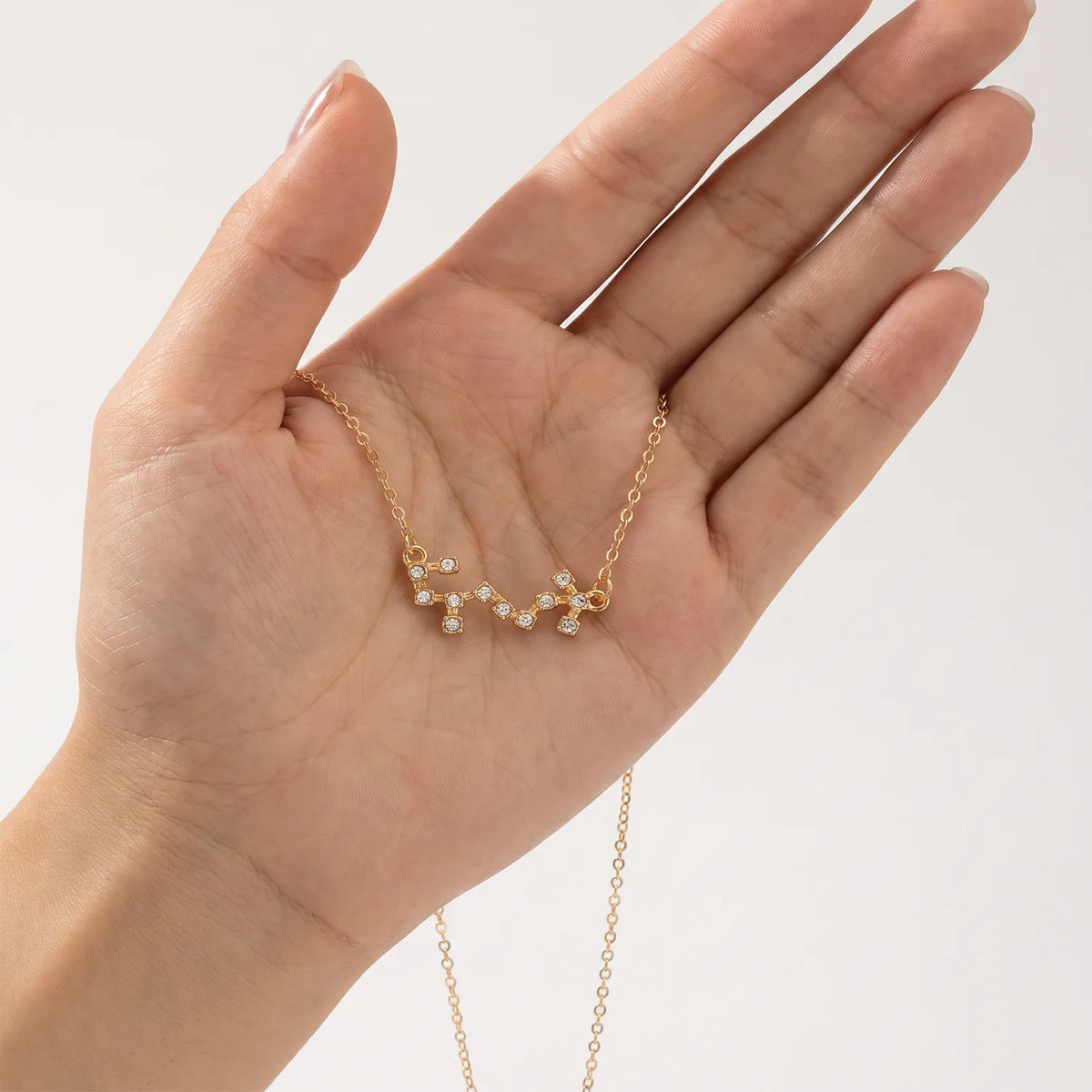 A delicate gold necklace with a scorpio constellation pendant set with tiny clear stones, held in the palm of a hand against a clean white background.