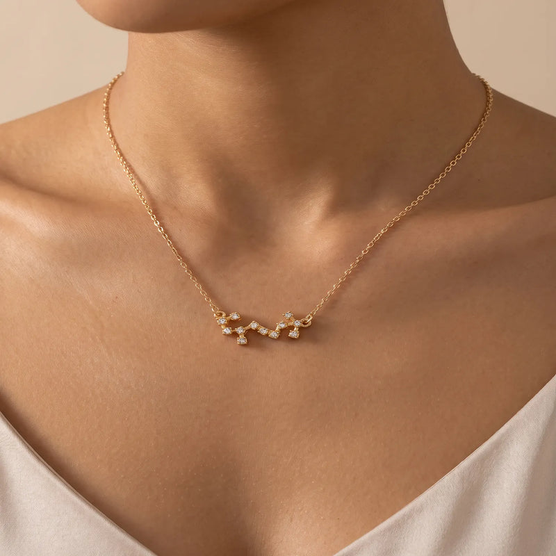 A close-up of a model's neck and upper chest wearing a delicate gold chain necklace with a constellation scorpio pendant set with tiny clear stones.