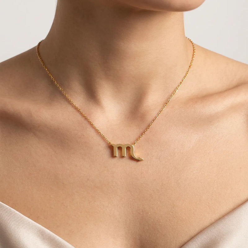 A close-up of a model's neck and chest wearing a delicate gold chain necklace with a zodiac sign Scorpio pendant.