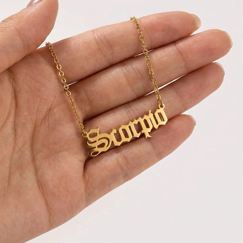 A gold nameplate necklace spelling scorpio in a scripted font held in a person's hand against a plain light background.