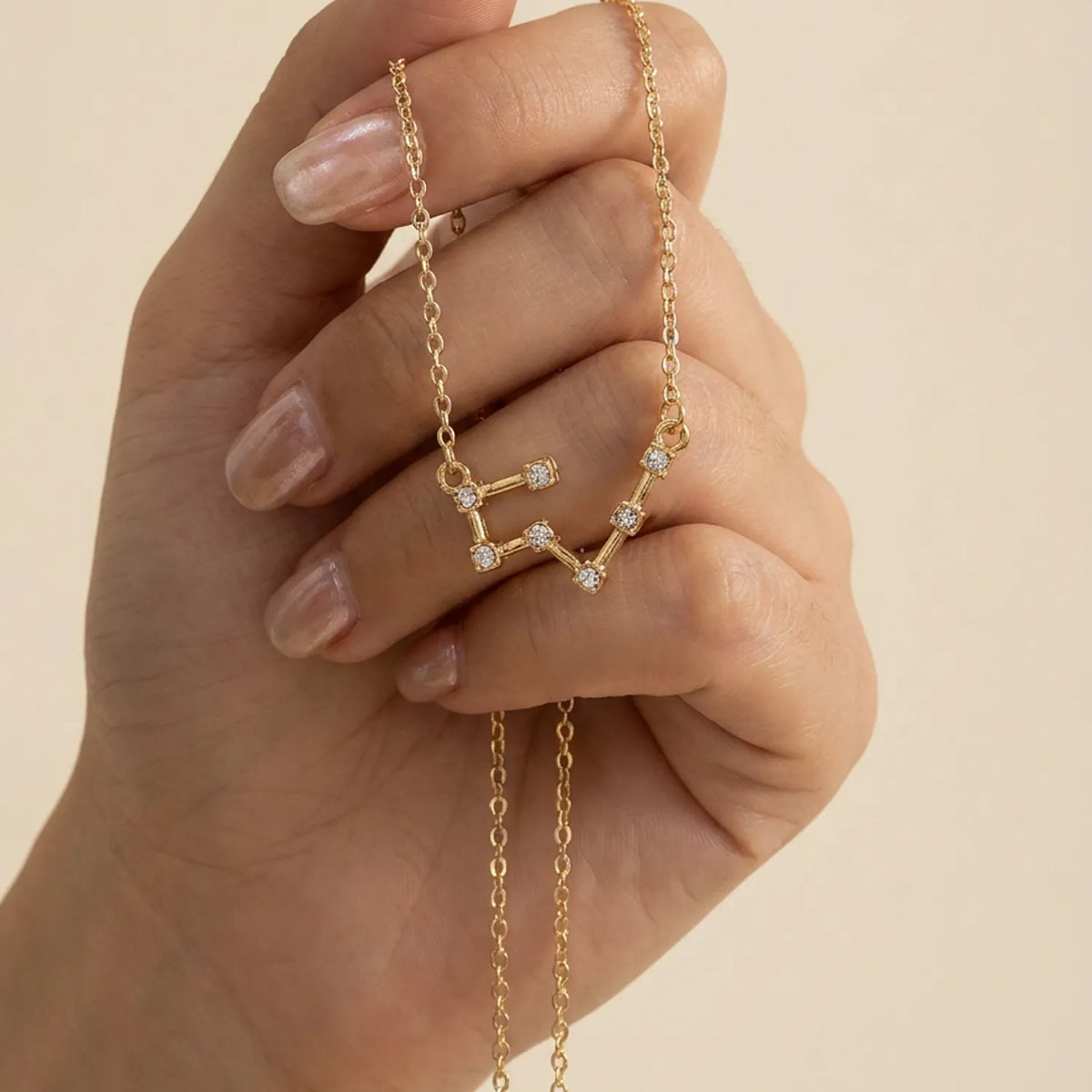 A gold chain necklace with a taurus constellation pendant, held in a hand with manicured nails against a warm beige background.