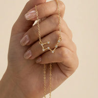 A gold chain necklace with a taurus constellation pendant, held in a hand with manicured nails against a warm beige background.