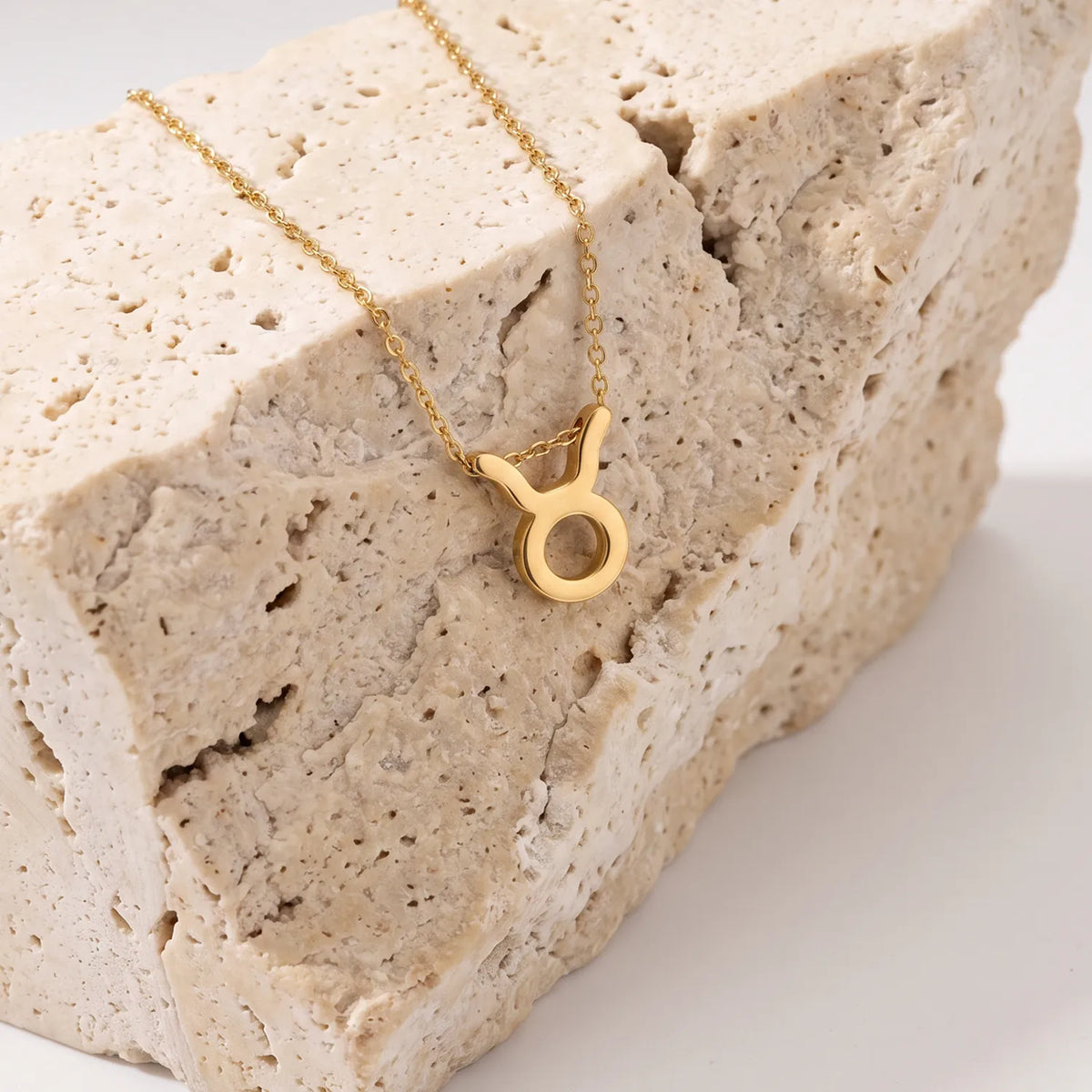 A gold necklace with a zodiac tausrus sign pendant, displayed on a textured beige stone block against a clean white background.