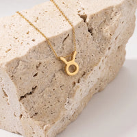 A gold necklace with a zodiac tausrus sign pendant, displayed on a textured beige stone block against a clean white background.