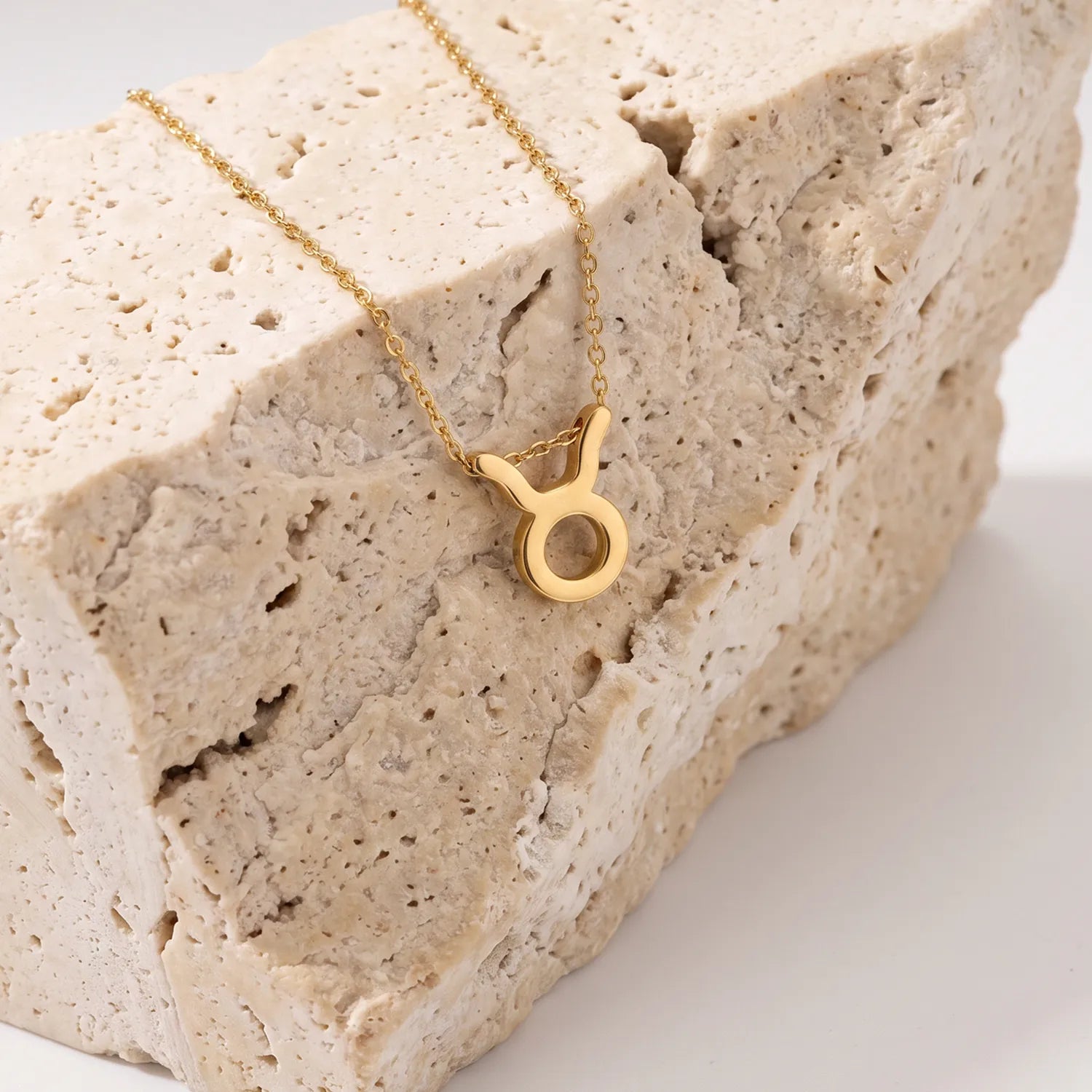A gold necklace with a zodiac tausrus sign pendant, displayed on a textured beige stone block against a clean white background.