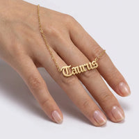 A gold nameplate necklace spelling 'Taurus' in a gothic font, worn on a person's ring finger. The delicate chain drapes across the back of the hand against a clean, light gray background.